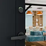 A wireless smart lock installed on an apartment door, with a furnished living room visible through the open doorway
