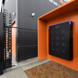 Black access control keypad reader in an orange metal mount outside a black building